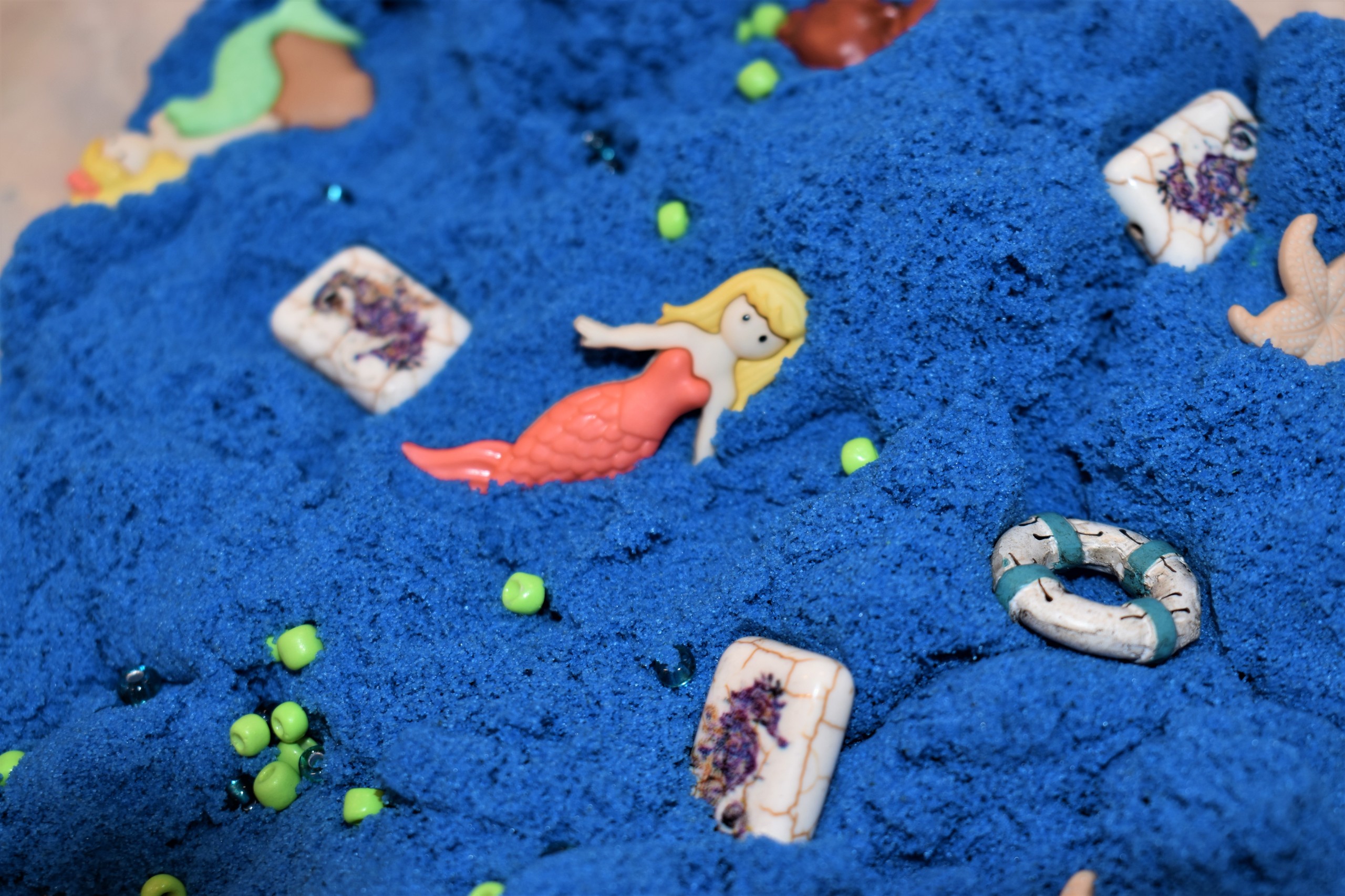 A plastic bin filled with blue kinetic sand has various beach themed objects inside.