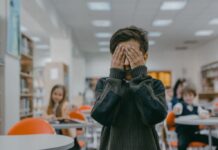 A boy covers his face with his hands against the backdrop of a classroom, indicating stress or anxiety.