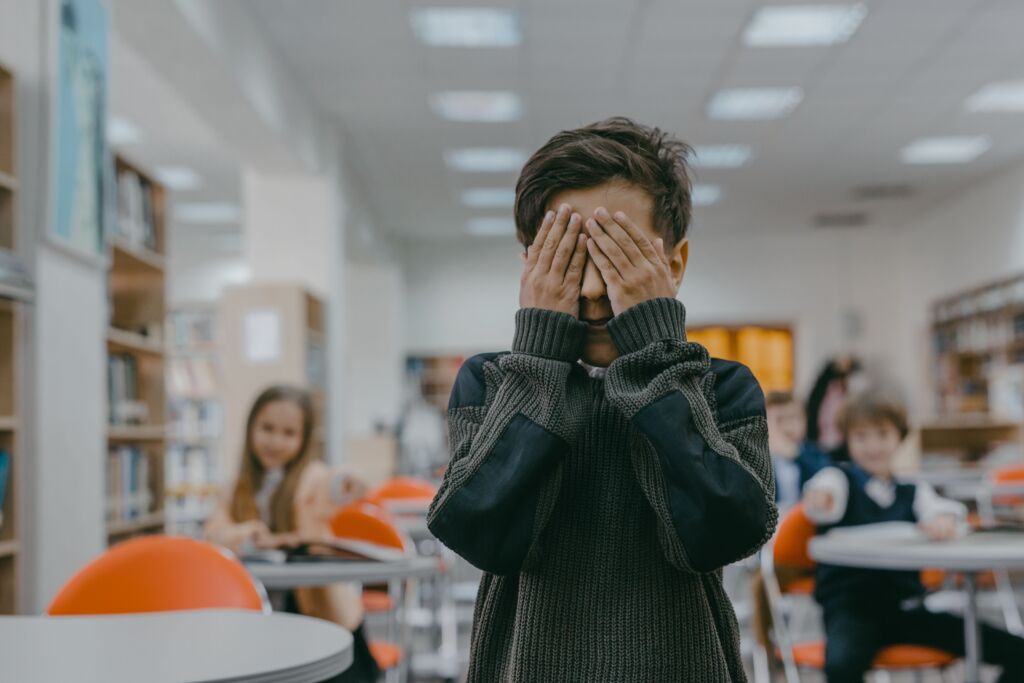 A boy covers his face with his hands against the backdrop of a classroom, indicating stress or anxiety.
