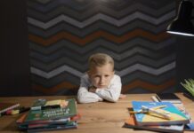 A boy crosses his arms at a table with school books, showing he is frustrated.