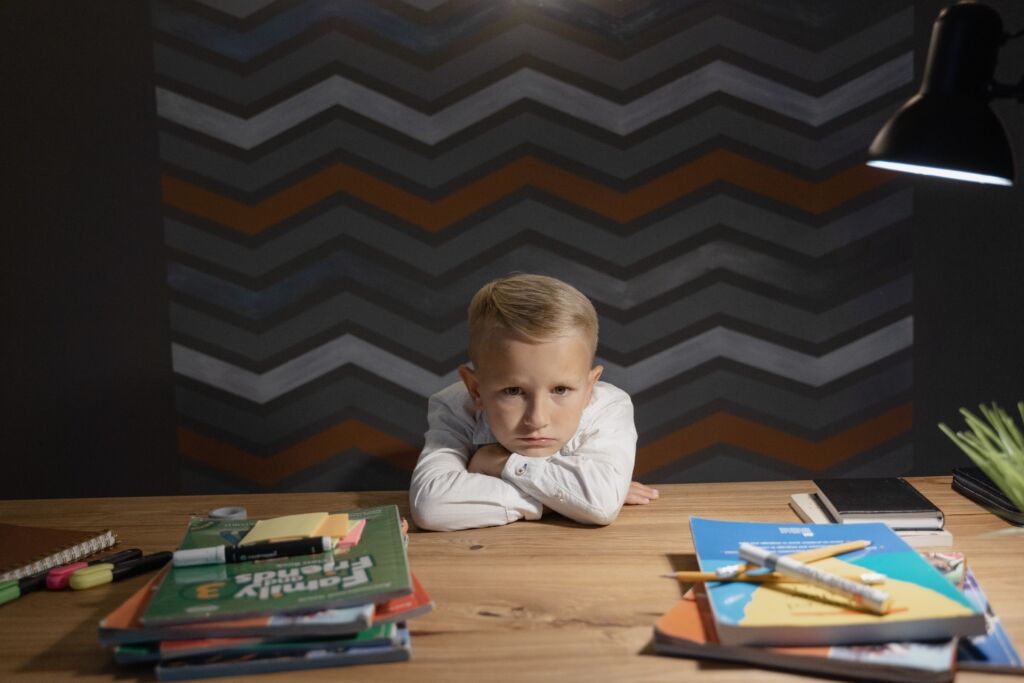 A boy crosses his arms at a table with school books, showing he is frustrated.