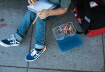 A kid sits on a sidewalk, with a chest-down view showing the child holding a notebook and pencil with a small box of other writing materials next to him.