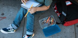 A kid sits on a sidewalk, with a chest-down view showing the child holding a notebook and pencil with a small box of other writing materials next to him.