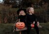 Stock photo of a blonde woman and a blonde girl sitting on a bench together next to a jack-o-lantern. They are dressed in all black and the woman is wearing a black pointed witch's hat. They are smiling.