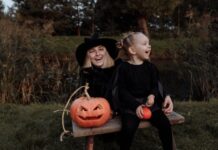 Stock photo of a blonde woman and a blonde girl sitting on a bench together next to a jack-o-lantern. They are dressed in all black and the woman is wearing a black pointed witch's hat. They are smiling.