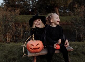 Stock photo of a blonde woman and a blonde girl sitting on a bench together next to a jack-o-lantern. They are dressed in all black and the woman is wearing a black pointed witch's hat. They are smiling.