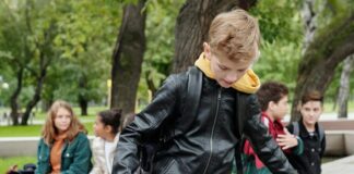 Stock photo of a boy using a skateboard at a park in front of a group of other tweens sitting on a ledge.