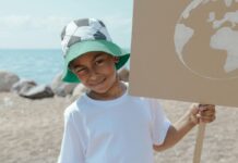 A child wearing a bucket hat with a soccer pattern on it squints one eye as he holds a cardboard sign with the Earth on it against a beach backdrop.