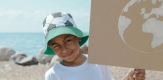 A child wearing a bucket hat with a soccer pattern on it squints one eye as he holds a cardboard sign with the Earth on it against a beach backdrop.
