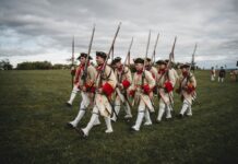 A group of men in traditional 18th-century uniforms reenact a historical march with rifles on a field.