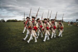 A group of men in traditional 18th-century uniforms reenact a historical march with rifles on a field.