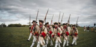 A group of men in traditional 18th-century uniforms reenact a historical march with rifles on a field.