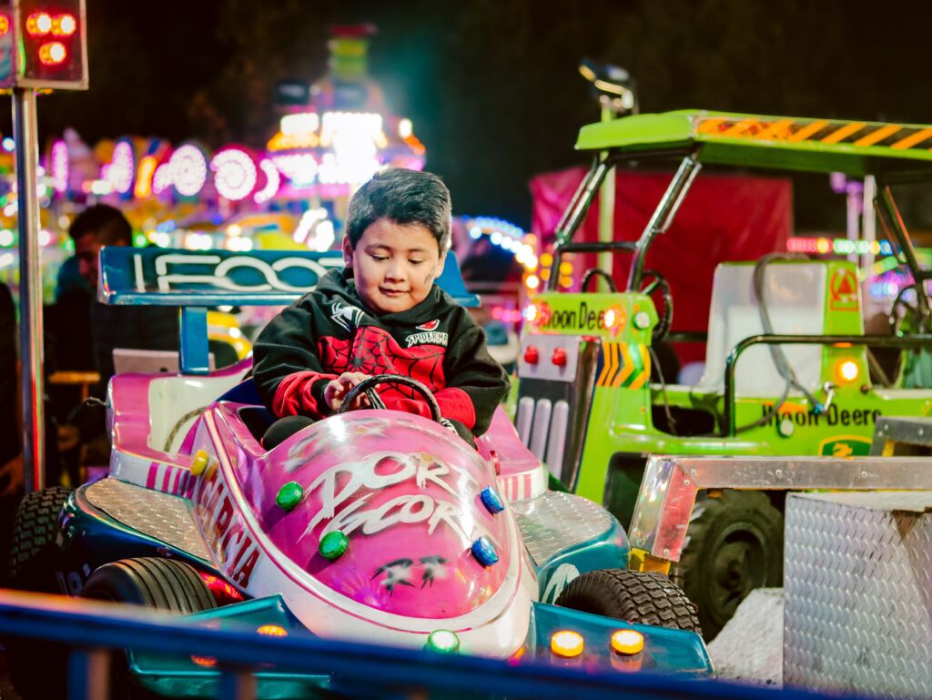 A boy sits in a pink ride car at a carnival at night with bright lights surrounding him in the background.